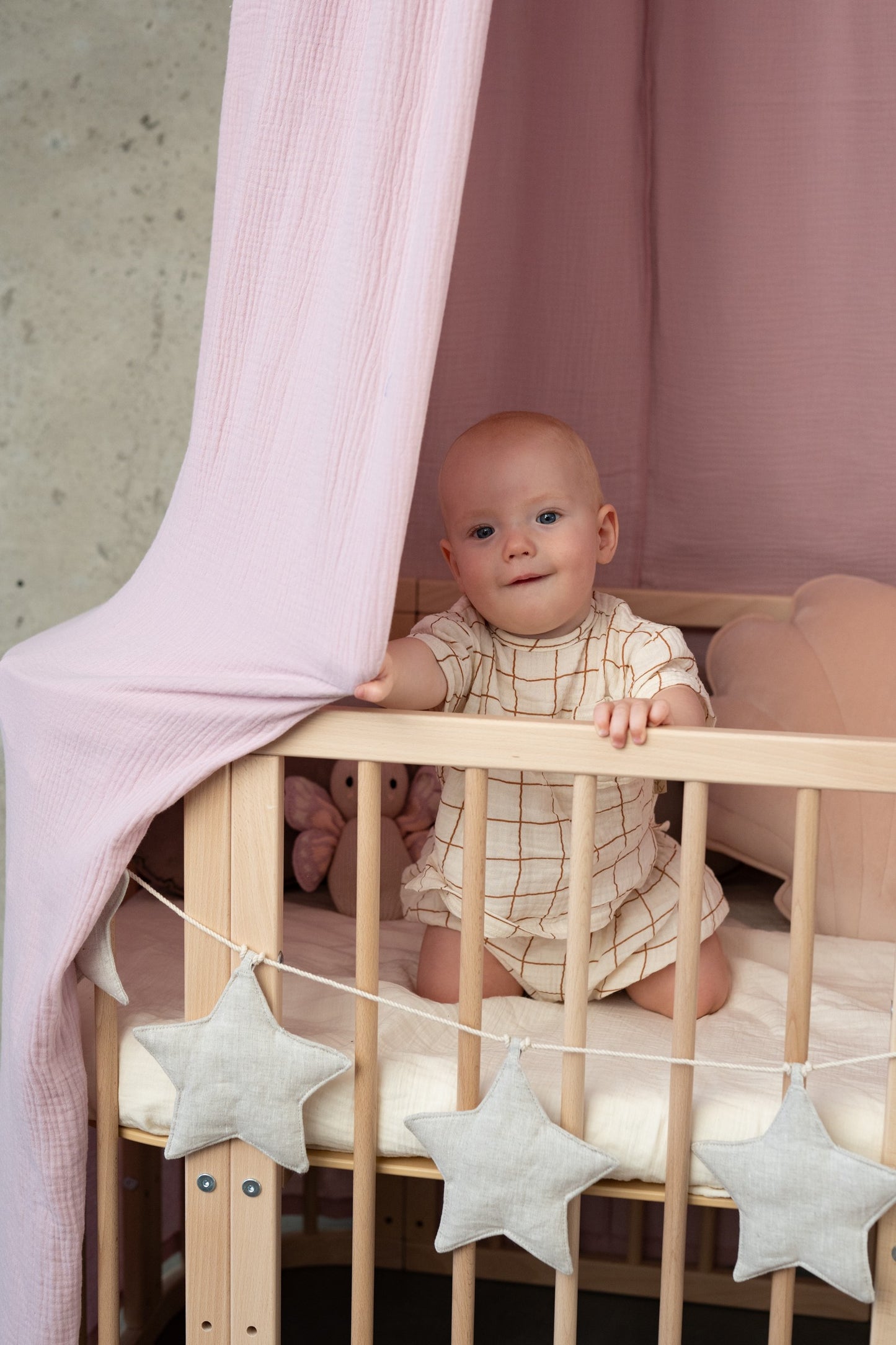 Baby playing in a crib, looking up at the soft hanging folds of a Moi Mili Baby Pink Organic Cotton Canopy in a bright nursery