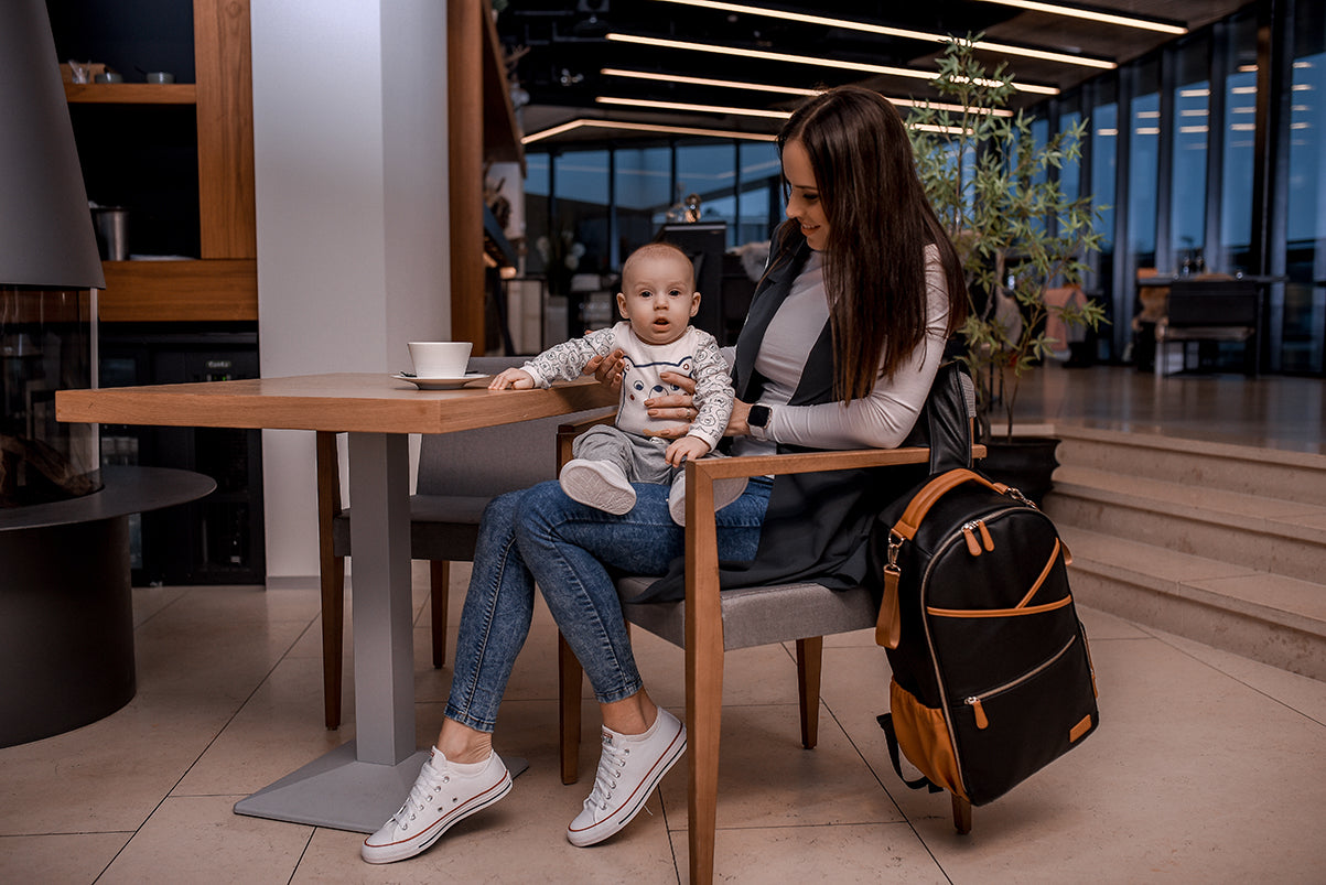 Lifestyle shot of a model with the Large Diaper Backpack – Black Coffee on a cafe table while holding her baby