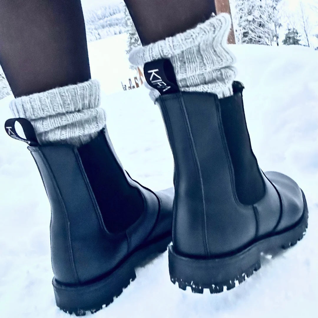 Black rubber boots with thick soles worn in the snow, paired with gray socks.