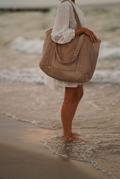 Moi Mili Mom Weekender Bag Latte: a mother standing in the ocean waves with a large cotton travel bag under a sunset sky