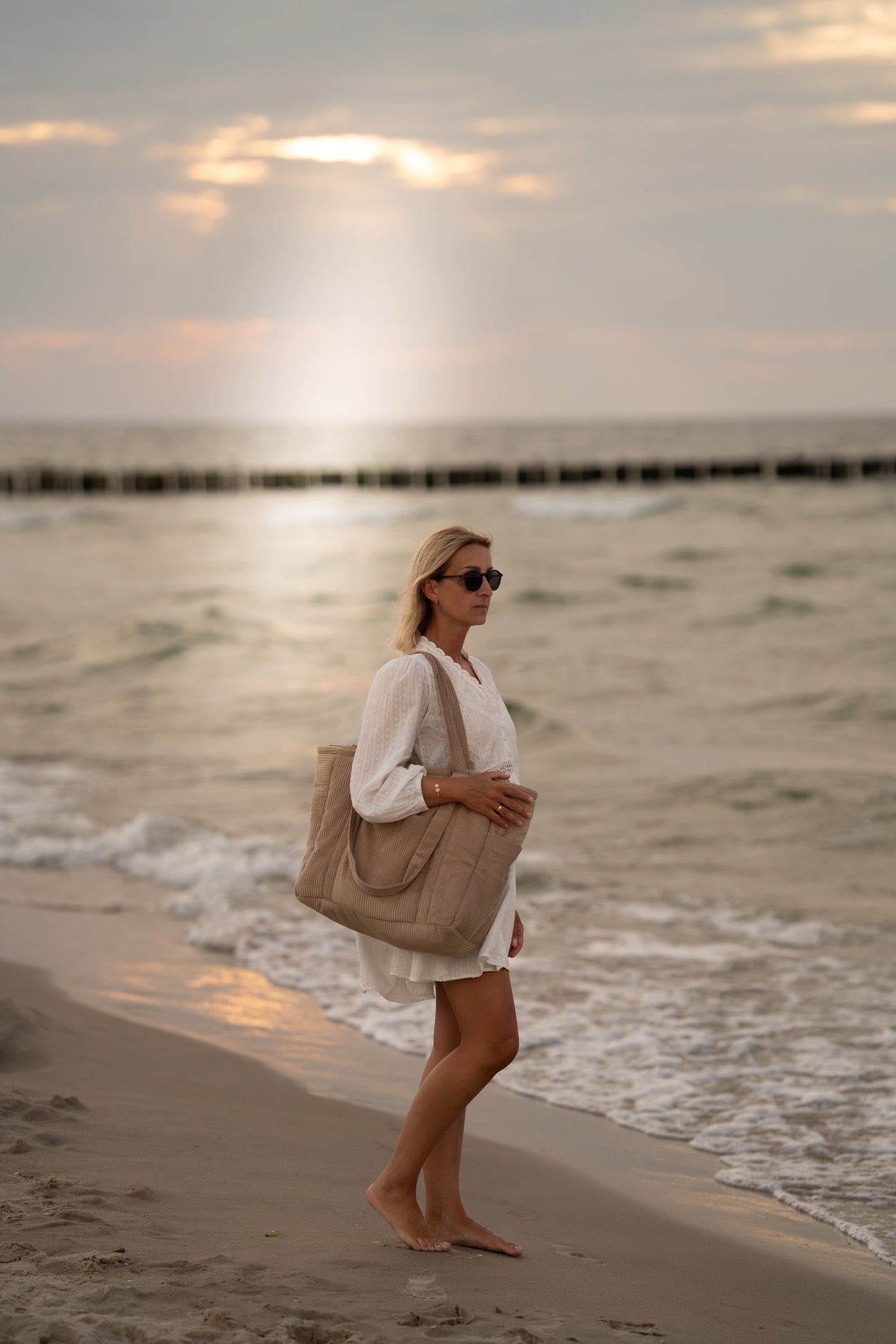 Moi Mili Mom Weekender Bag Latte: a mother walking along the shore with a sturdy quilted bag on her shoulder at sunset