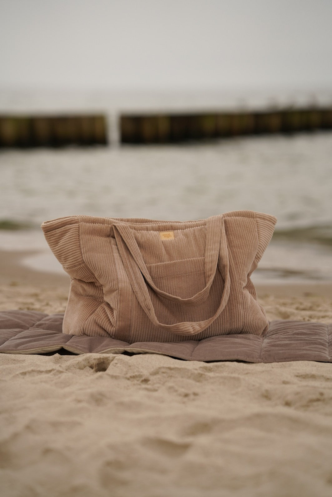 Moi Mili Mom Weekender Bag Latte: a large quilted cotton travel bag resting on the sand at a quiet beach