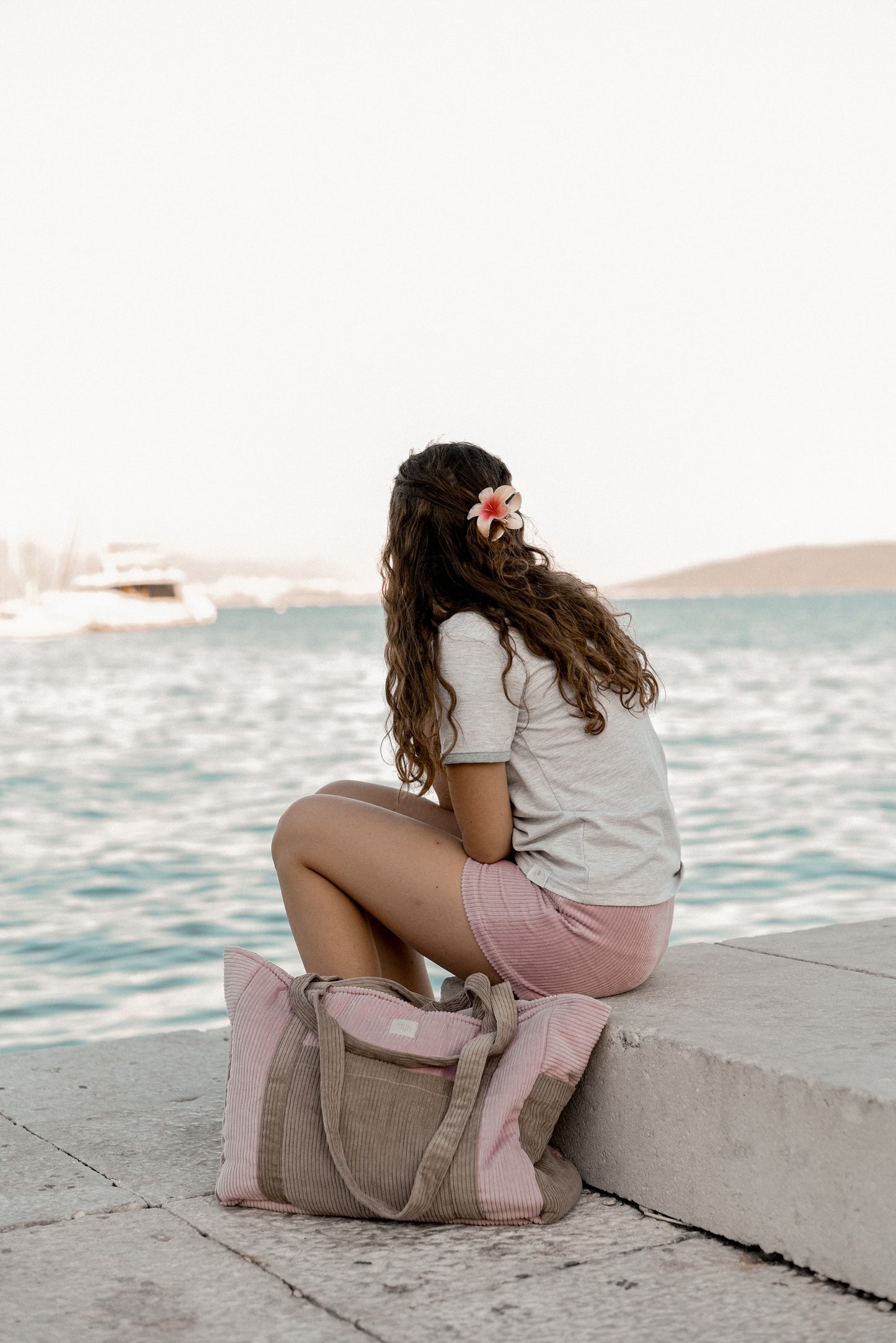 Moi Mili Mom Weekender Bag Pink Coffee: back view of a young woman sitting on a dock with her large quilted cotton bag beside her at the waterfront