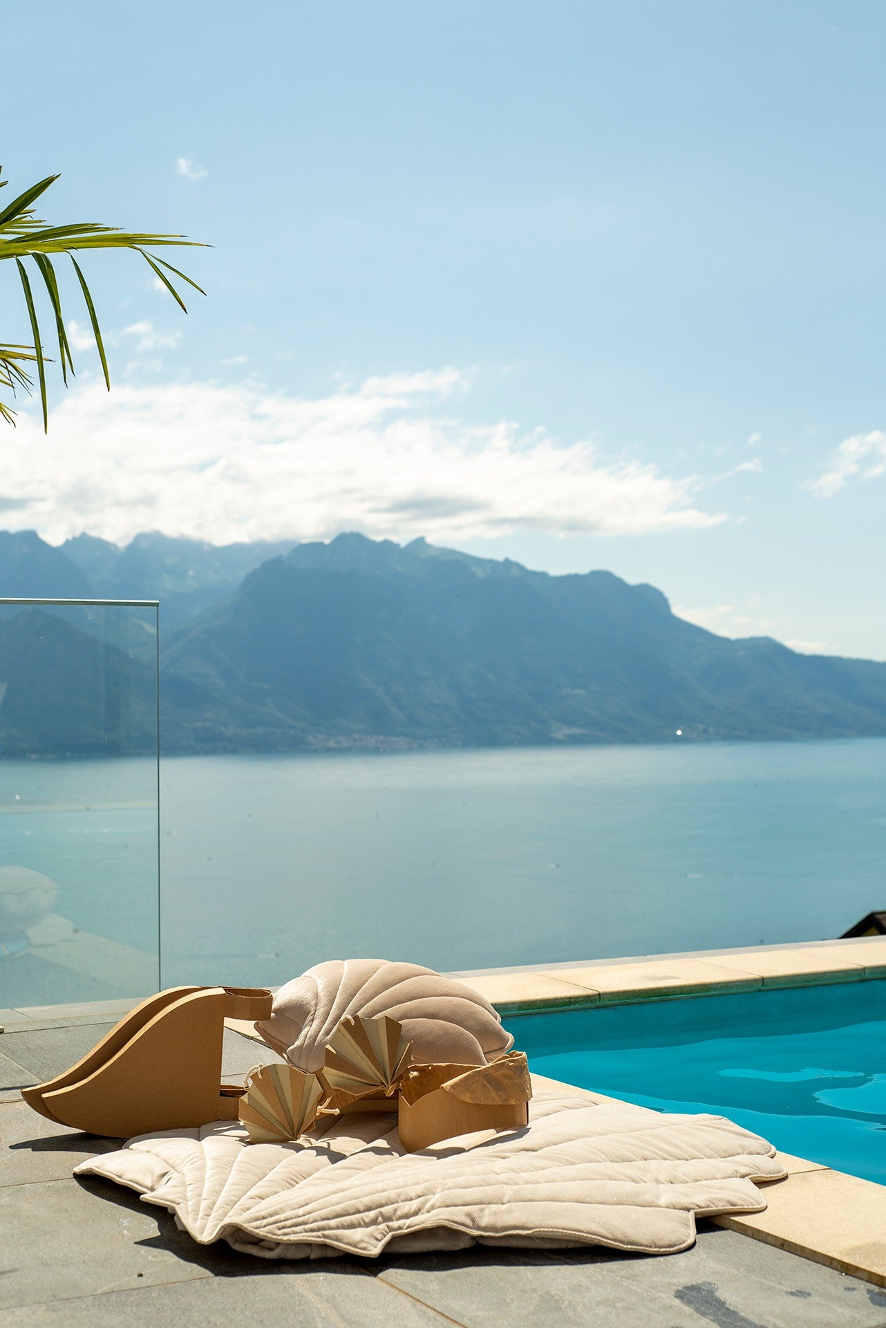 Moi Mili Beige Shell Playmat: a luxurious velvet shell-shaped baby mat resting at a poolside with a stunning sea and mountain view background
