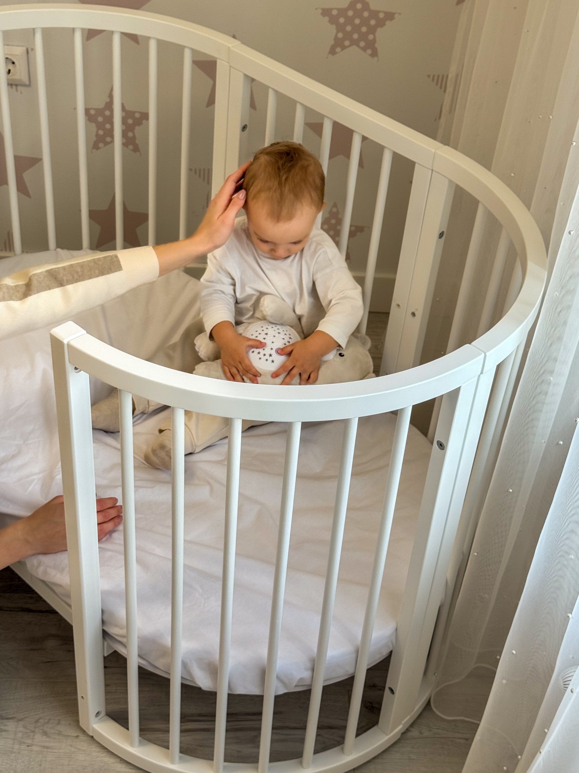 baby  sitting inside the Round Baby Crib OLIN with Mattress - White playing with a soft toy