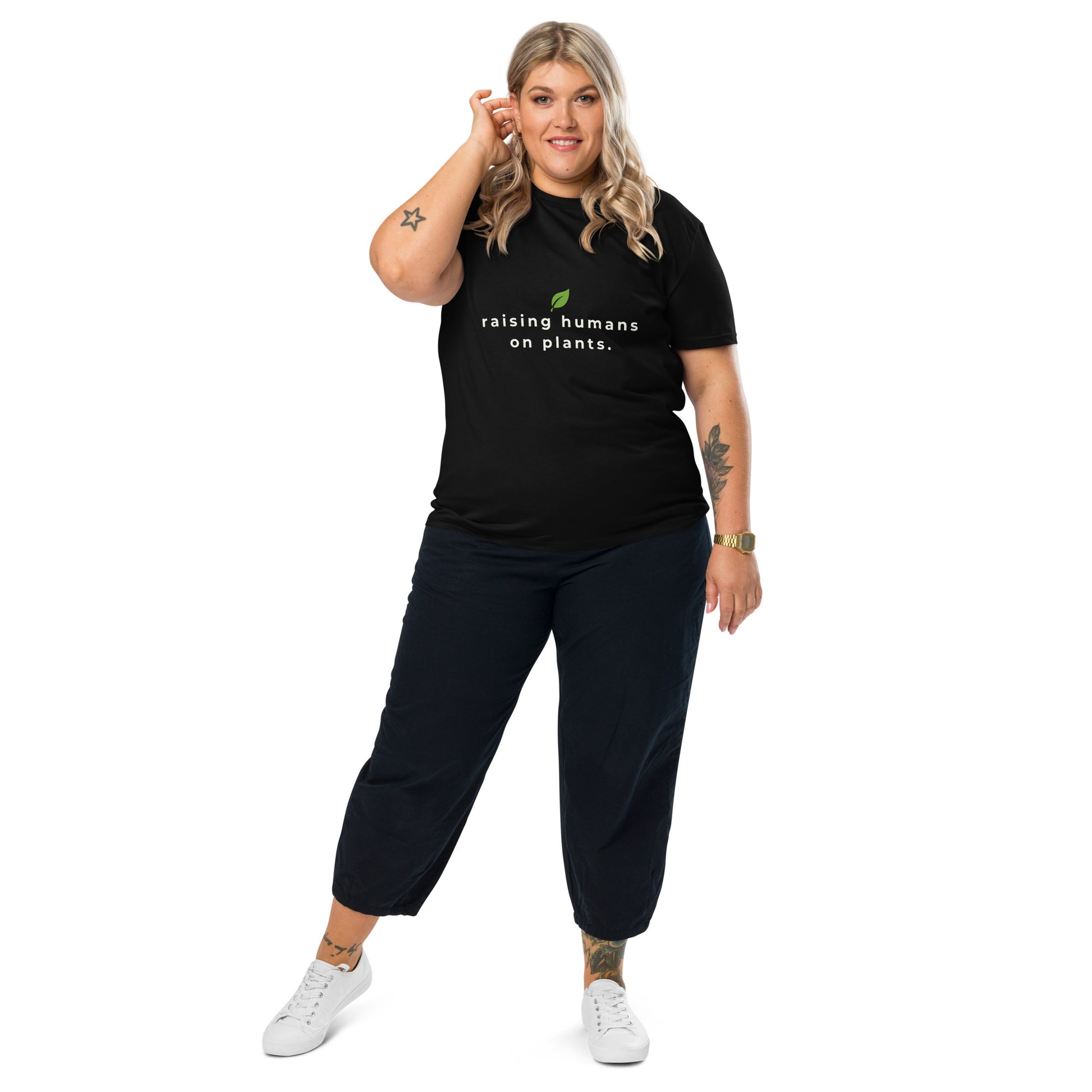Black plant-based women's t-shirt worn by a model, featuring the quote "raising humans on plants." with a decorative leaf on top.