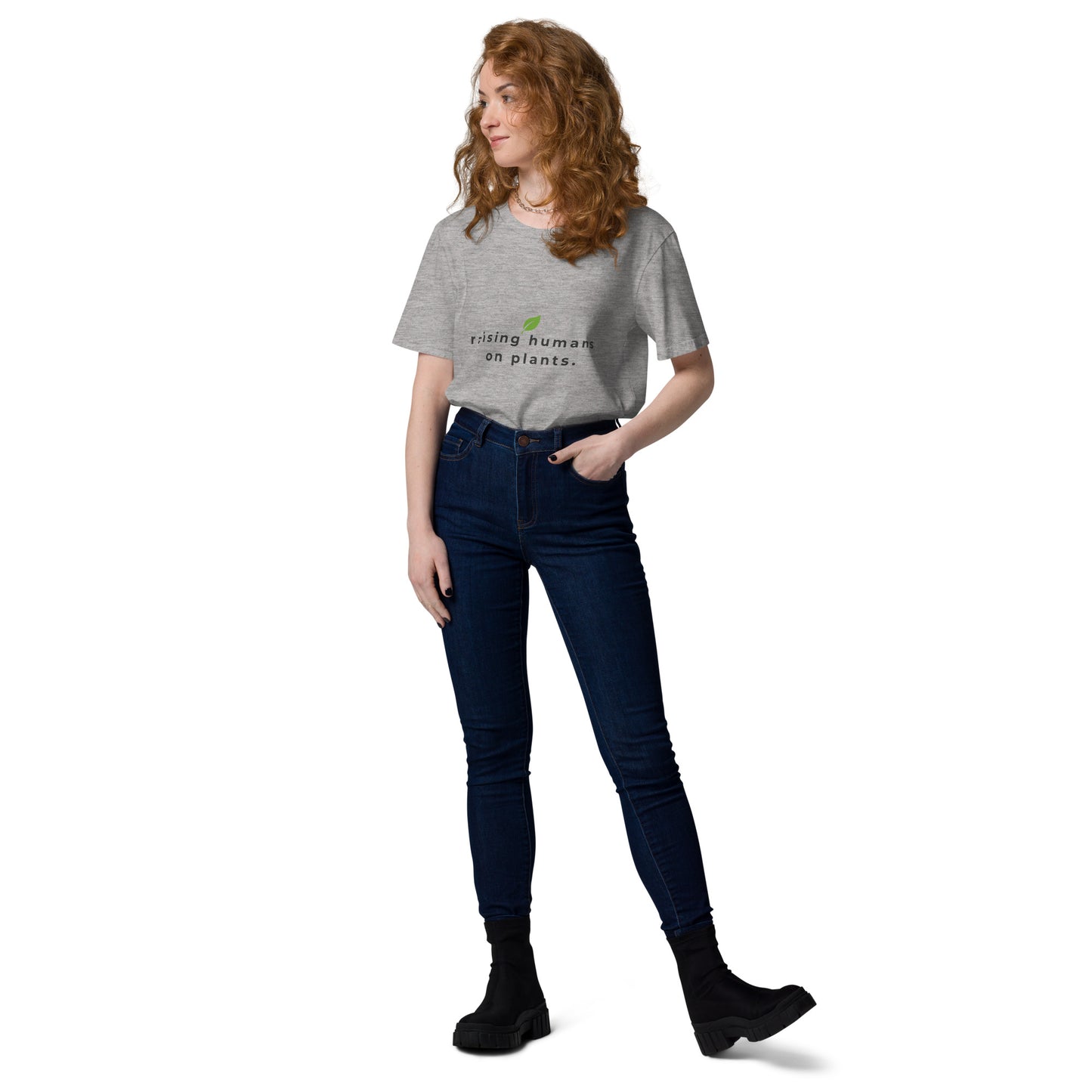 Woman posing casually in a heather grey organic cotton t-shirt. Featuring the minimalist text "raising humans on plants." with a green leaf logo design.