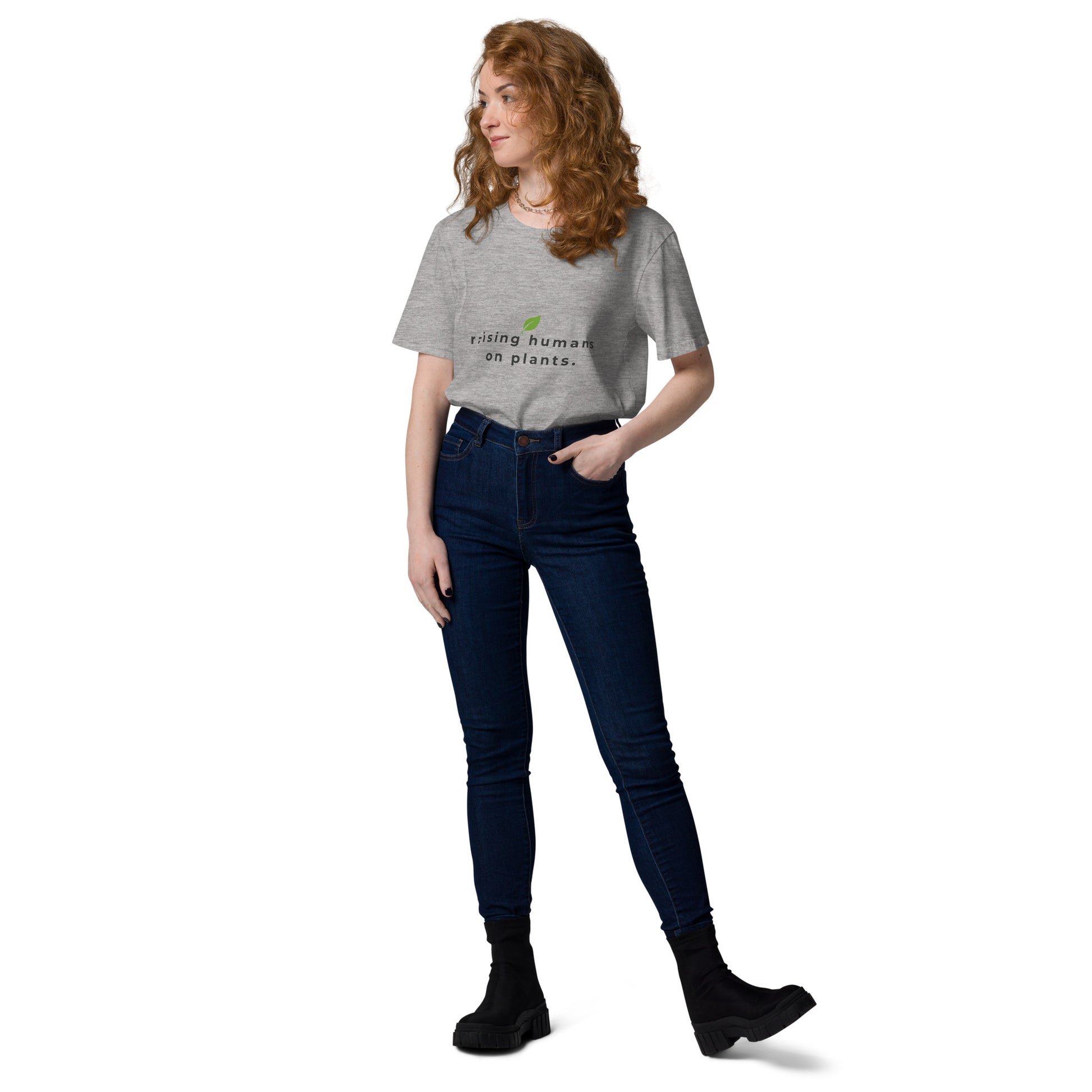 Woman posing casually in a heather grey organic cotton t-shirt. Featuring the minimalist text "raising humans on plants." with a green leaf logo design.