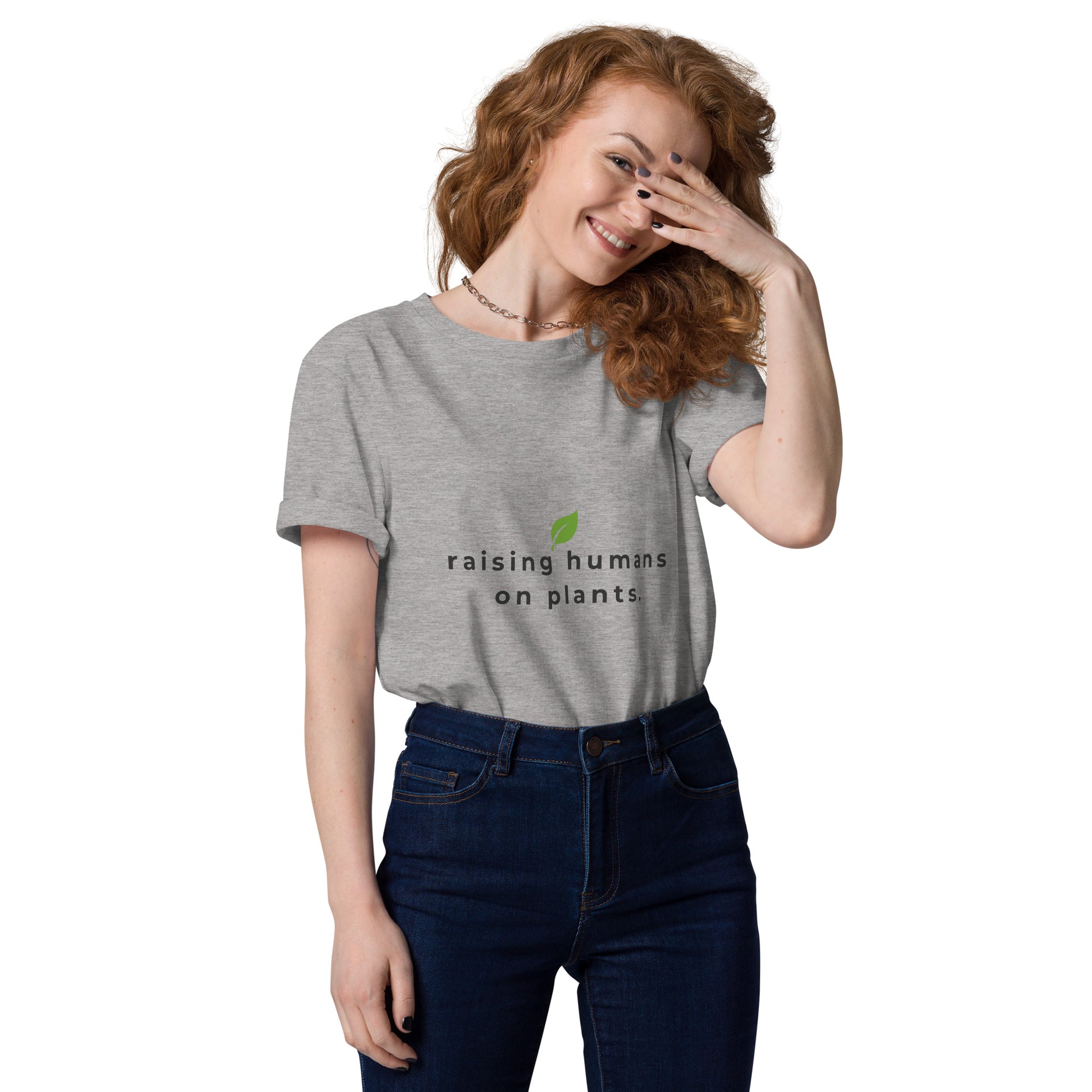 Smiling model wearing an eco-friendly grey t-shirt. On the chest is printed the phrase "raising humans on plants." in charcoal grey with a small leaf centered above the text.